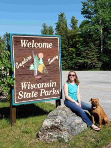 Young woman sits on a large rock in front of a sign that reads Welcome to Wisconsin State Parks. A dog sits at her feet.