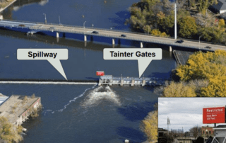 Aerial view of Menasha Dam on Lake Winnebago, showing labeled spillway and tainter gates. Water flows over the spillway while the tainter gates control outflow.