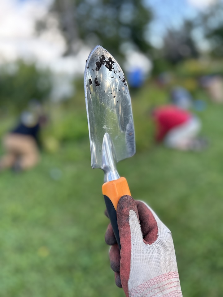 A gloved hand holding a gardening trowel with dirt on the blade, with volunteers working in the background during a shoreline volunteer day.