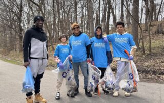 Five volunteers pose on a wooded trail holding full bags of collected trash during the 2025 Fox-Wolf Watershed Cleanup. All wear blue event shirts and gloves.