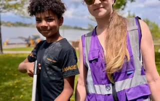 A young boy and woman stand smiling in a sunny park near the water. The boy holds a trash grabber tool, and the woman wears a purple reflective vest and sunglasses. Trees and the Fox River are visible in the background.