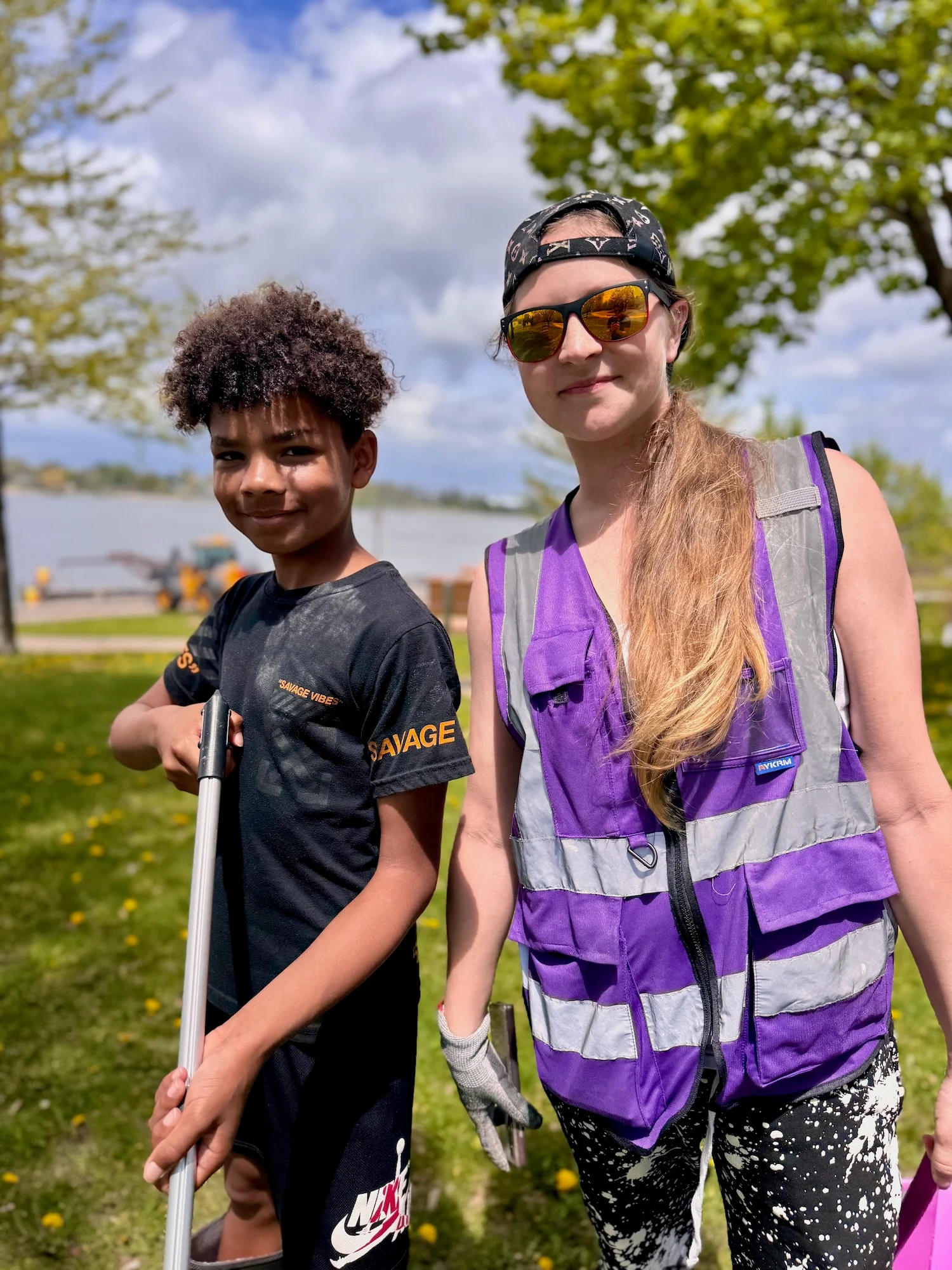 A young boy and woman stand smiling in a sunny park near the water. The boy holds a trash grabber tool, and the woman wears a purple reflective vest and sunglasses. Trees and the Fox River are visible in the background.