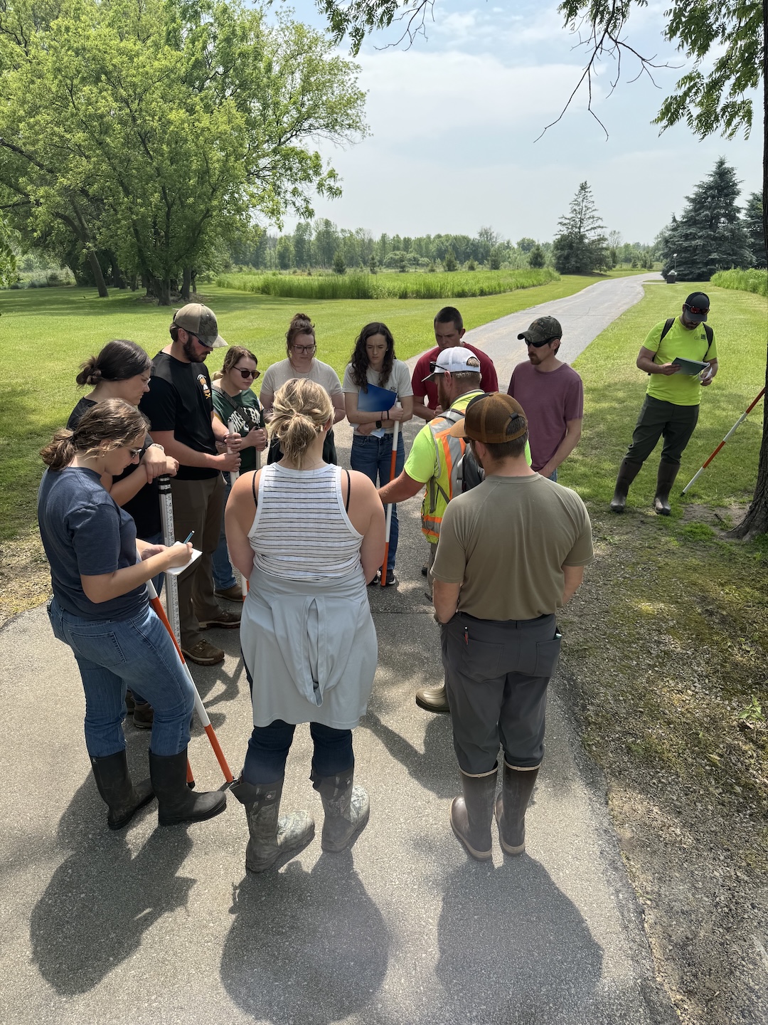 A group of conservation professionals in boots and outdoor gear stand in a circle on a paved path, listening to instructions during a field training on streambank erosion.