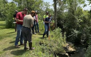 A group of people stands near a stream, using measuring tools to assess erosion along the bank. Lush greenery and trees surround the scene on a sunny day.