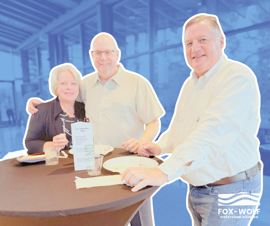 Three guests enjoy food and conversation at the 2024 Happiest Hour, hosted by the Fox-Wolf Watershed Alliance. They stand around a tall table featuring a prompt card encouraging discussions about clean water.