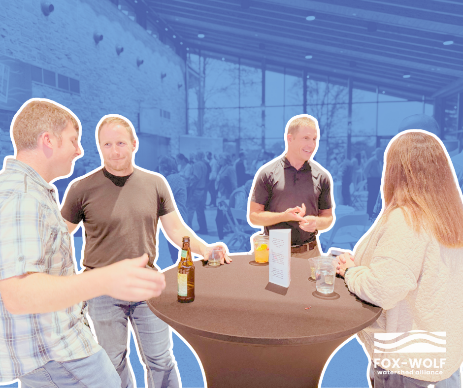 Four attendees share drinks and laughter during the 2024 Happiest Hour. They’re gathered at a cocktail table with a clean water conversation card, surrounded by a bustling, blue-tinted event venue.