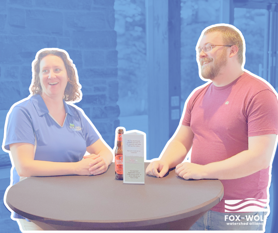 A woman in a Fox-Wolf Watershed Alliance polo chats with a guest at the 2024 Happiest Hour. They smile across a small table with a clean water conversation prompt and a bottle of hard cider.