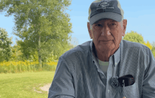 Chuck Farrey seated outdoors in Winnebago County, looking toward the camera with a field and trees behind him.
