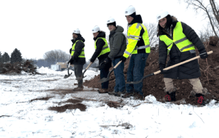 Five project partners in hard hats and high-visibility vests stand in a snowy construction area, each holding a shovel during the Brothertown Creek restoration groundbreaking. A pile of soil and winter trees are visible behind them.