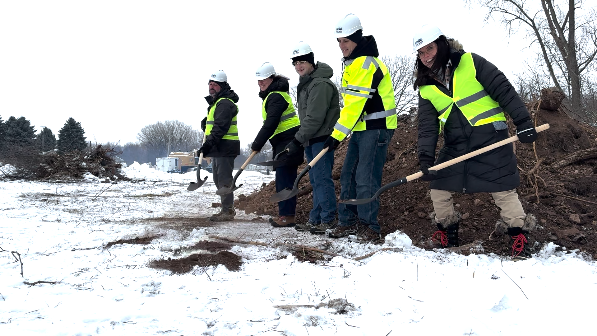 Five project partners in hard hats and high-visibility vests stand in a snowy construction area, each holding a shovel during the Brothertown Creek restoration groundbreaking. A pile of soil and winter trees are visible behind them.