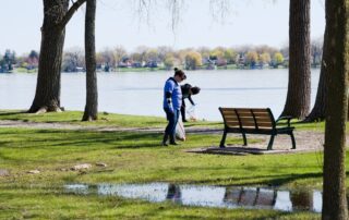 Two volunteers pick up trash along a grassy lakeshore, with trees, a park bench, and a puddle in the foreground on a clear spring day.