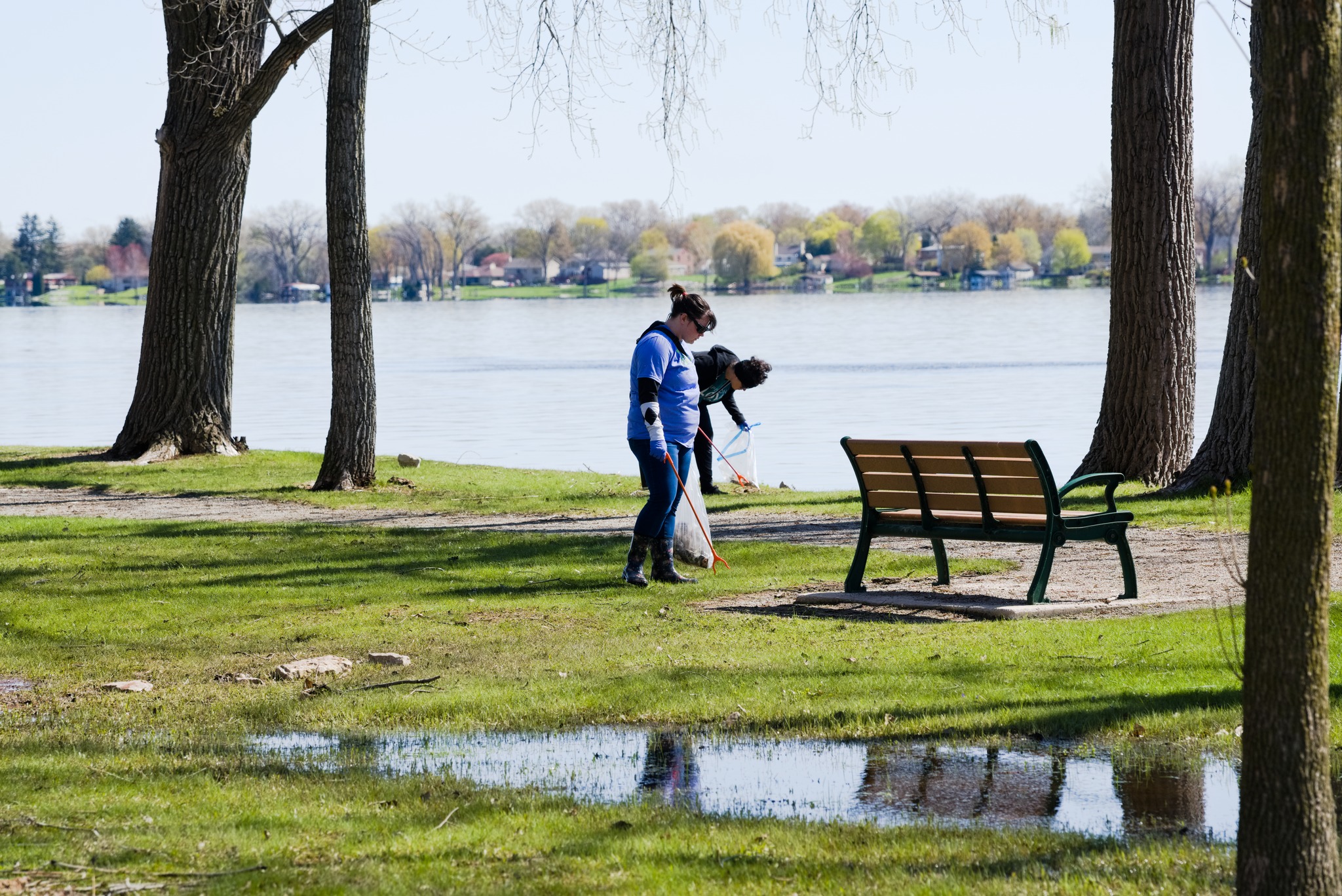 Two volunteers pick up trash along a grassy lakeshore, with trees, a park bench, and a puddle in the foreground on a clear spring day.