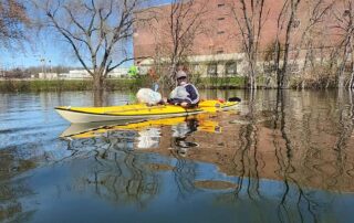 A volunteer in a yellow kayak paddles along a calm urban stretch of the Fox River, a bag of collected trash resting across the bow, with bare trees and a brick building reflected in the water.