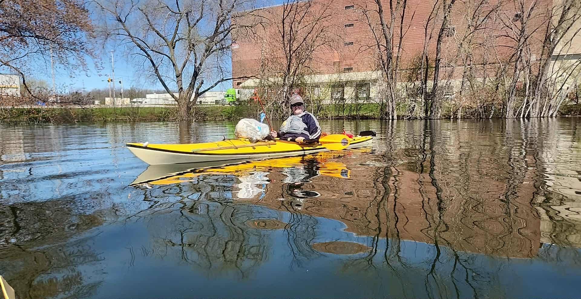 A volunteer in a yellow kayak paddles along a calm urban stretch of the Fox River, a bag of collected trash resting across the bow, with bare trees and a brick building reflected in the water.