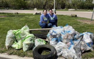 Two smiling volunteers crouch behind a large collection of filled trash bags, a wooden board, and a tire pulled from the watershed during the 2026 Fox-Wolf Annual Cleanup.