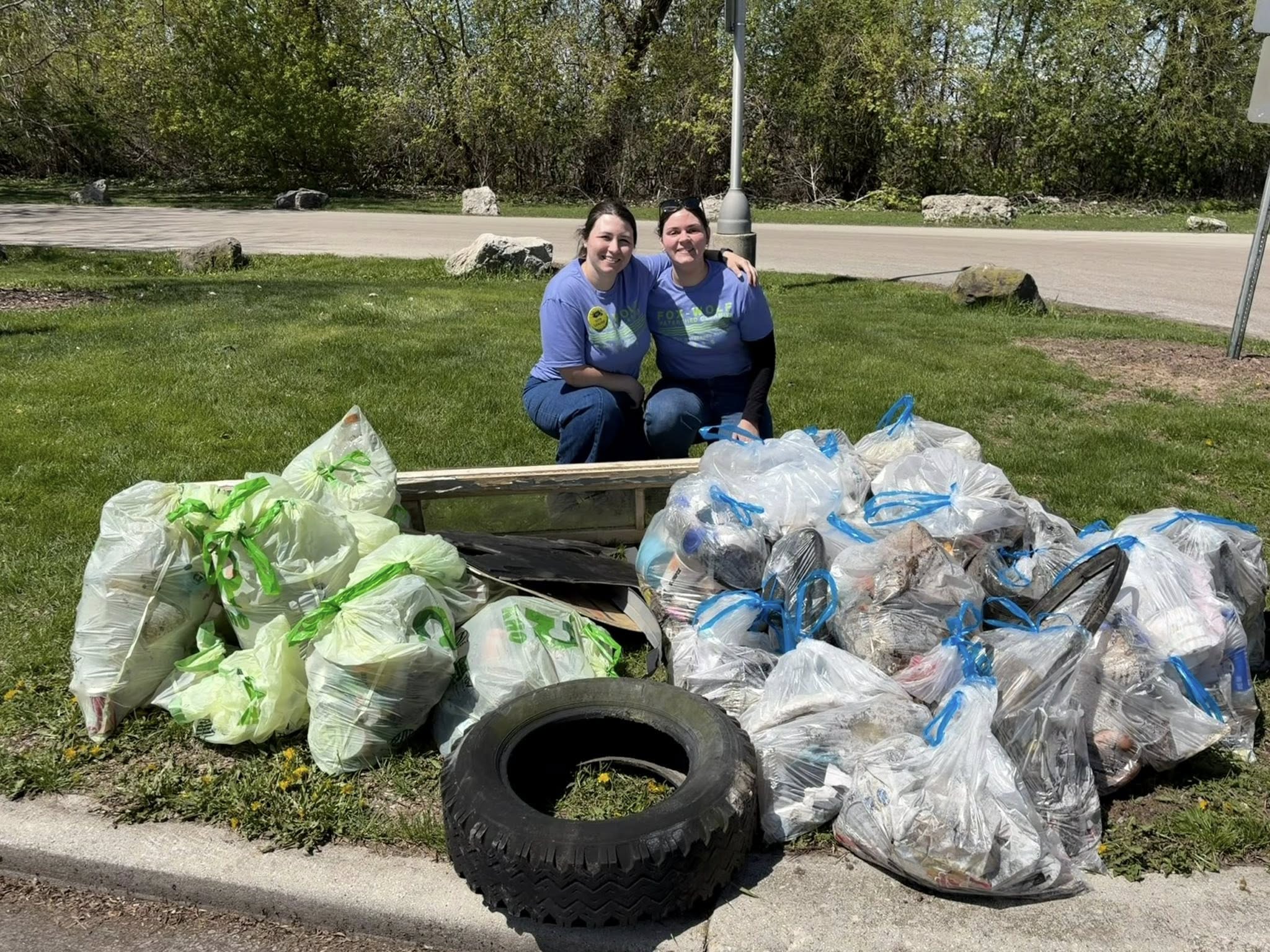 Two smiling volunteers crouch behind a large collection of filled trash bags, a wooden board, and a tire pulled from the watershed during the 2026 Fox-Wolf Annual Cleanup.