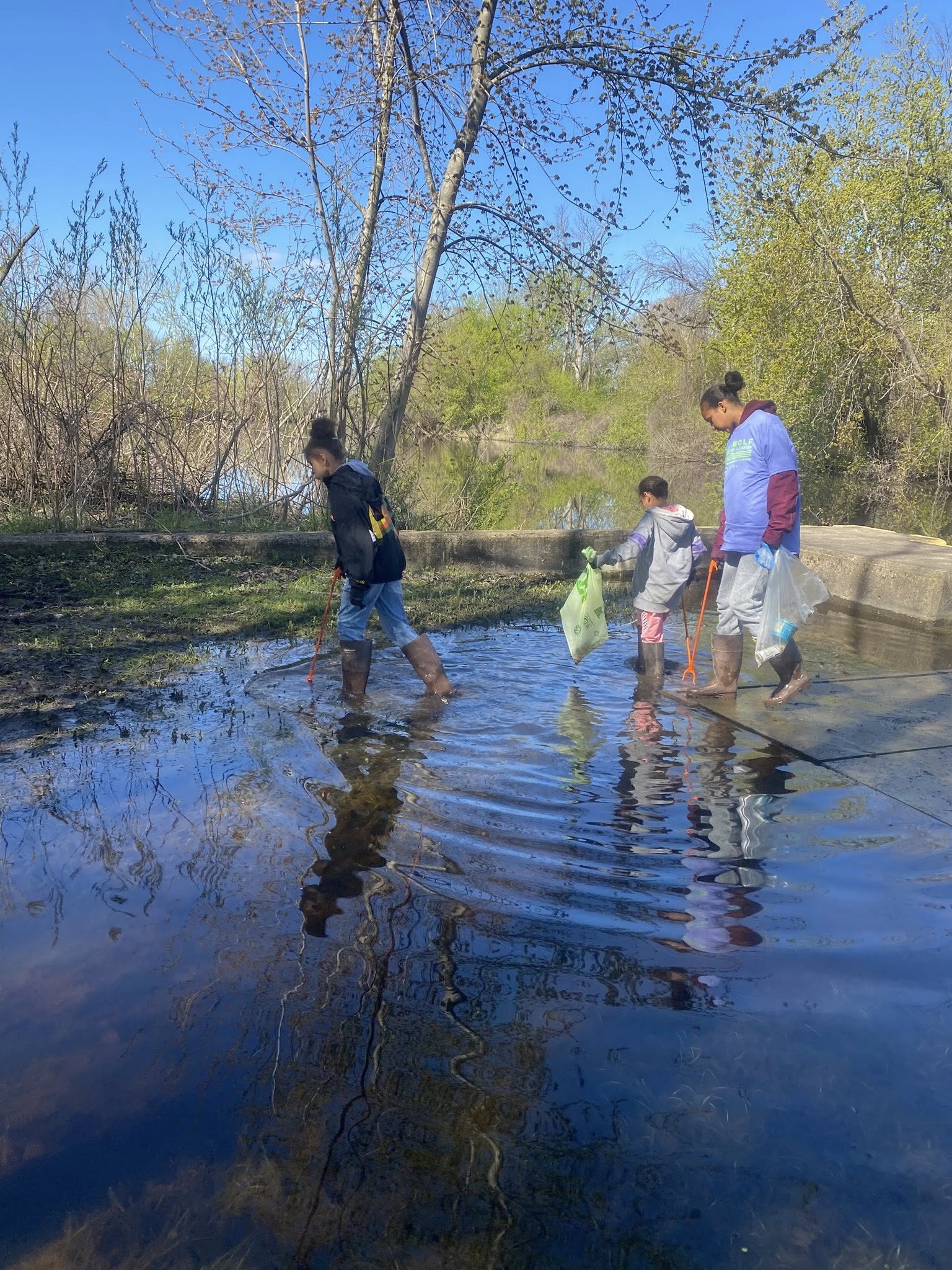Three volunteers, including a child, wade through a shallow stretch of water along a wooded riverbank, collecting trash on a sunny spring day.